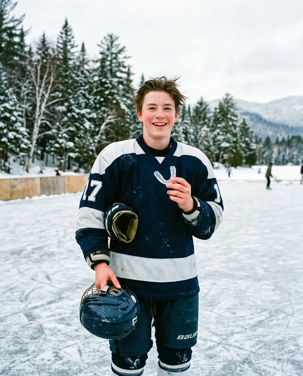 A smiling teenage hockey player in a navy and white number 17 jersey stands on an outdoor rink ringed by snowy evergreens, holding up a clear mouthguard, with a glove tucked under one arm and his helmet in his other hand.